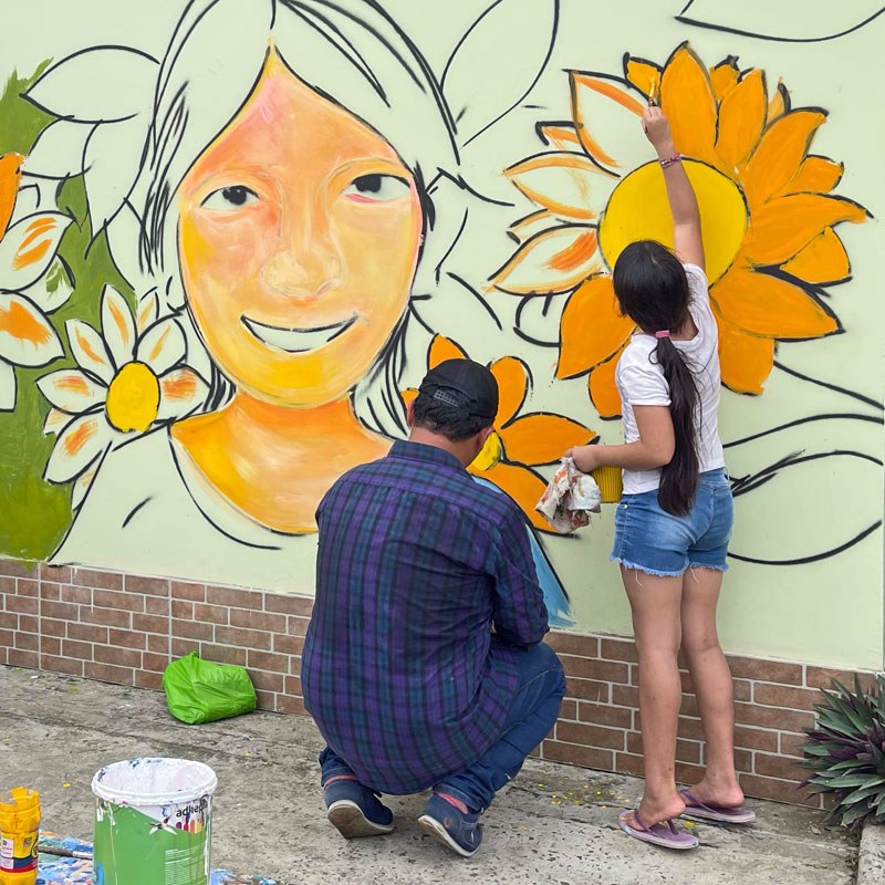 Mural comunitario — Fundación Visión Raíces Un hombre y una joven pintan juntos un mural de girasoles y un retrato en una pared comunitaria, actividad artística de la Fundación Visión Raíces