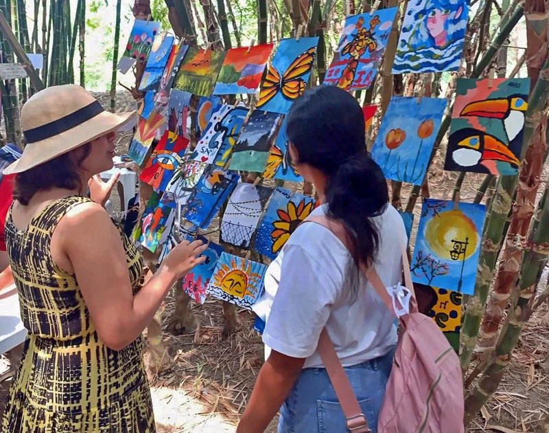 Dos mujeres observan y comentan una exhibición de pinturas infantiles colgadas entre bambú durante el evento Slow Food, Fundación Visión Raíces