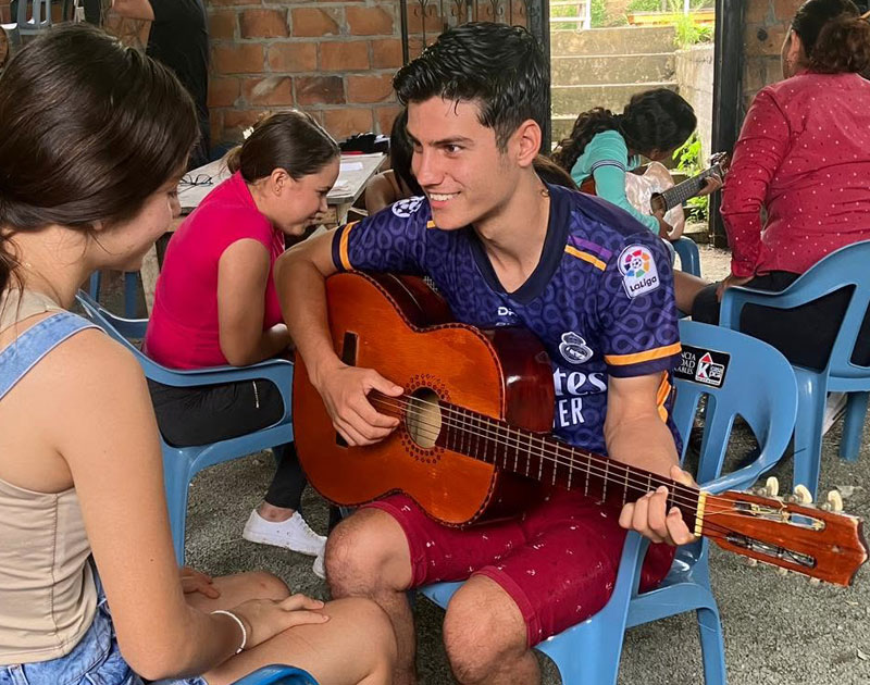 Joven toca guitarra sonriendo junto a otros participantes sentados en sillas al aire libre, durante un taller de música del proyecto Finaroma, Fundación Visión Raíces