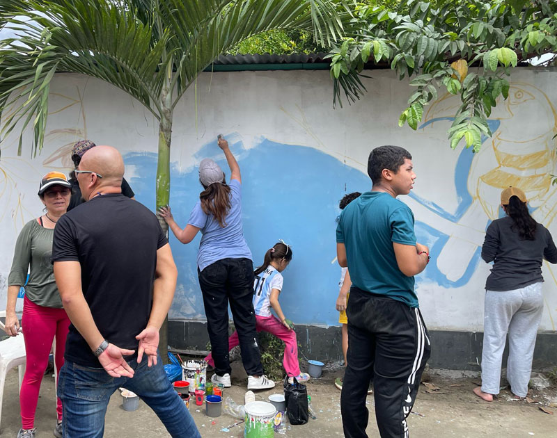 Adultos y niños pintan juntos la base azul de un mural comunitario al aire libre, con el boceto de una figura ya trazado sobre la pared, Fundación Visión Raíces