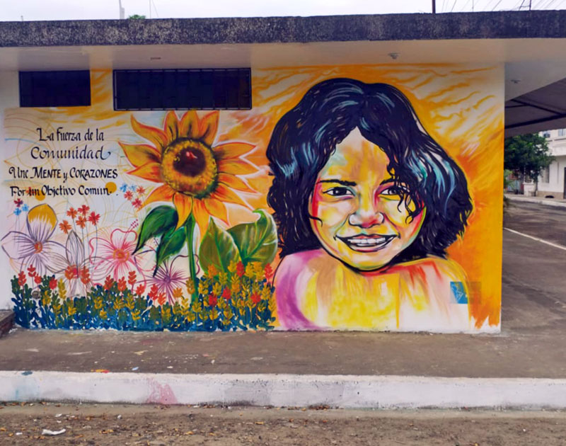 Mural terminado con el retrato de una niña sonriente, un girasol y el mensaje "La fuerza de la comunidad une mentes y corazones por un objetivo común", Fundación Visión Raíces