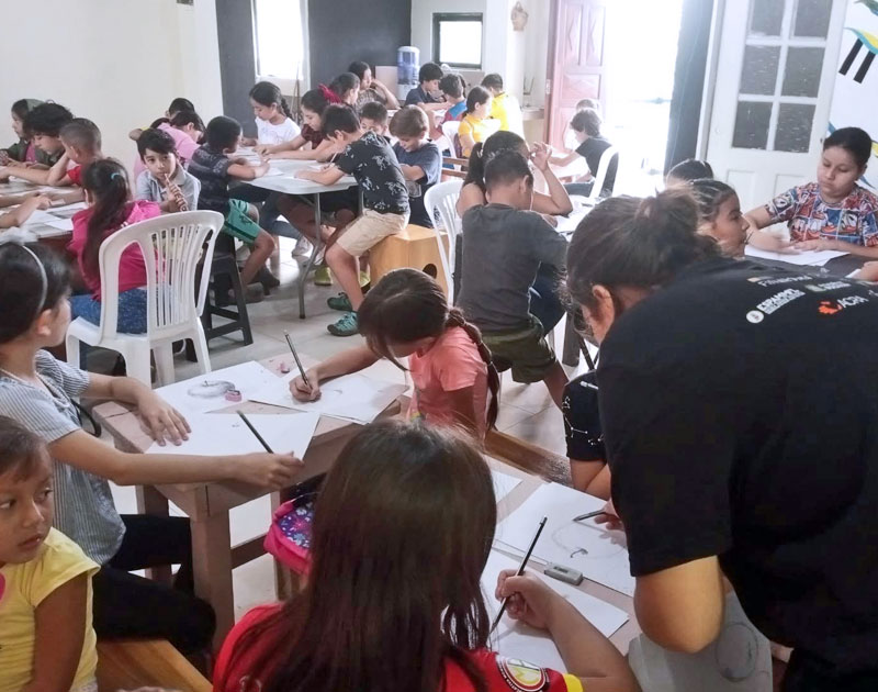 Grupo numeroso de niños dibujando en mesas dentro de un salón comunitario, con un facilitador guiando el taller, proyecto Finaroma de la Fundación Visión Raíces